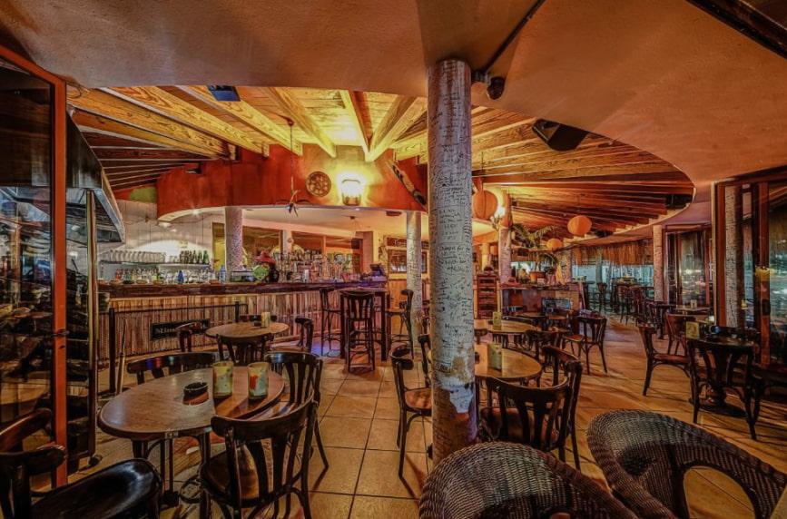 Waikiki Cocktail Bar Vista general nocturna del interior del bar, con mesas altas de madera y decoración ecléctica